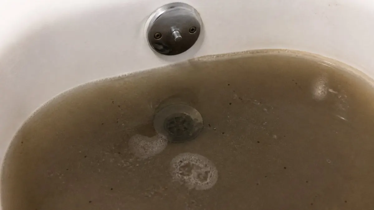 Close-up of a residential bathtub drain with dark, murky water pooling around it, bathroom lighting overhead, water line visible against white porcelain