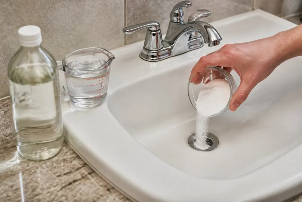 Close-up of a hand pouring baking soda into a white bathroom sink drain, with a bottle of white vinegar and a measuring cup visible on the counter beside the sink under warm bathroom lighting