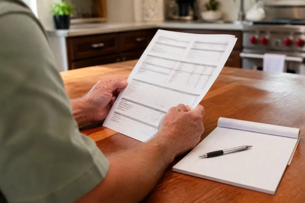Close-up of hands holding and reviewing a detailed written HVAC estimate document on kitchen table with pen and notepad, natural lighting